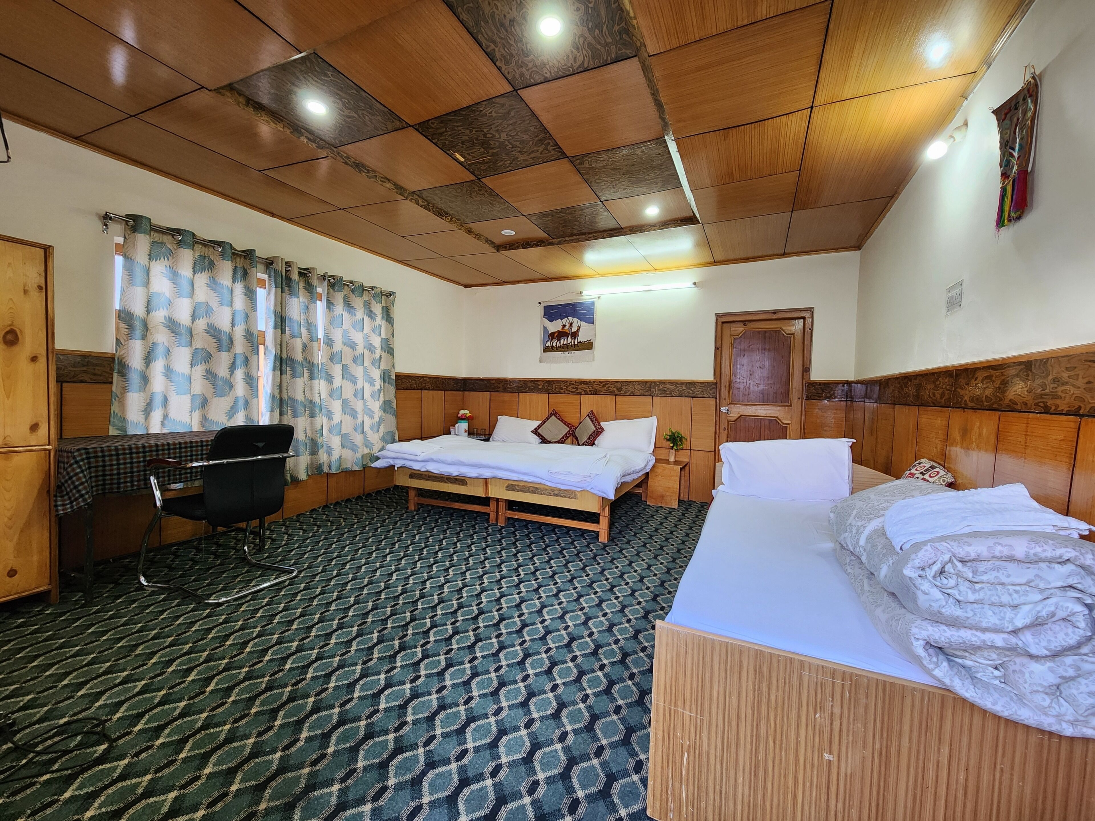 Une chambre d’hôtel avec un plafond en bois, un lit recouvert de draps blancs, un bureau avec une chaise et une moquette à motifs.