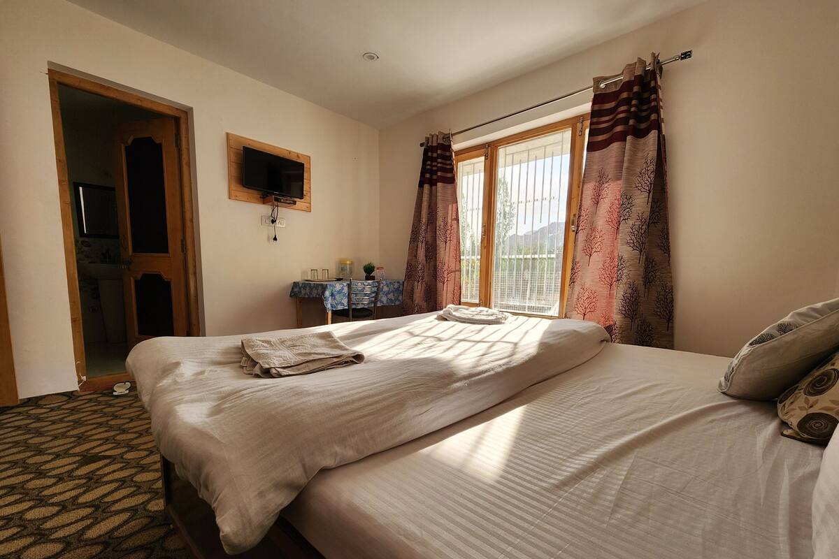 A bedroom with a bed, a TV mounted on the wall, a desk with a chair, and a window with curtains.
