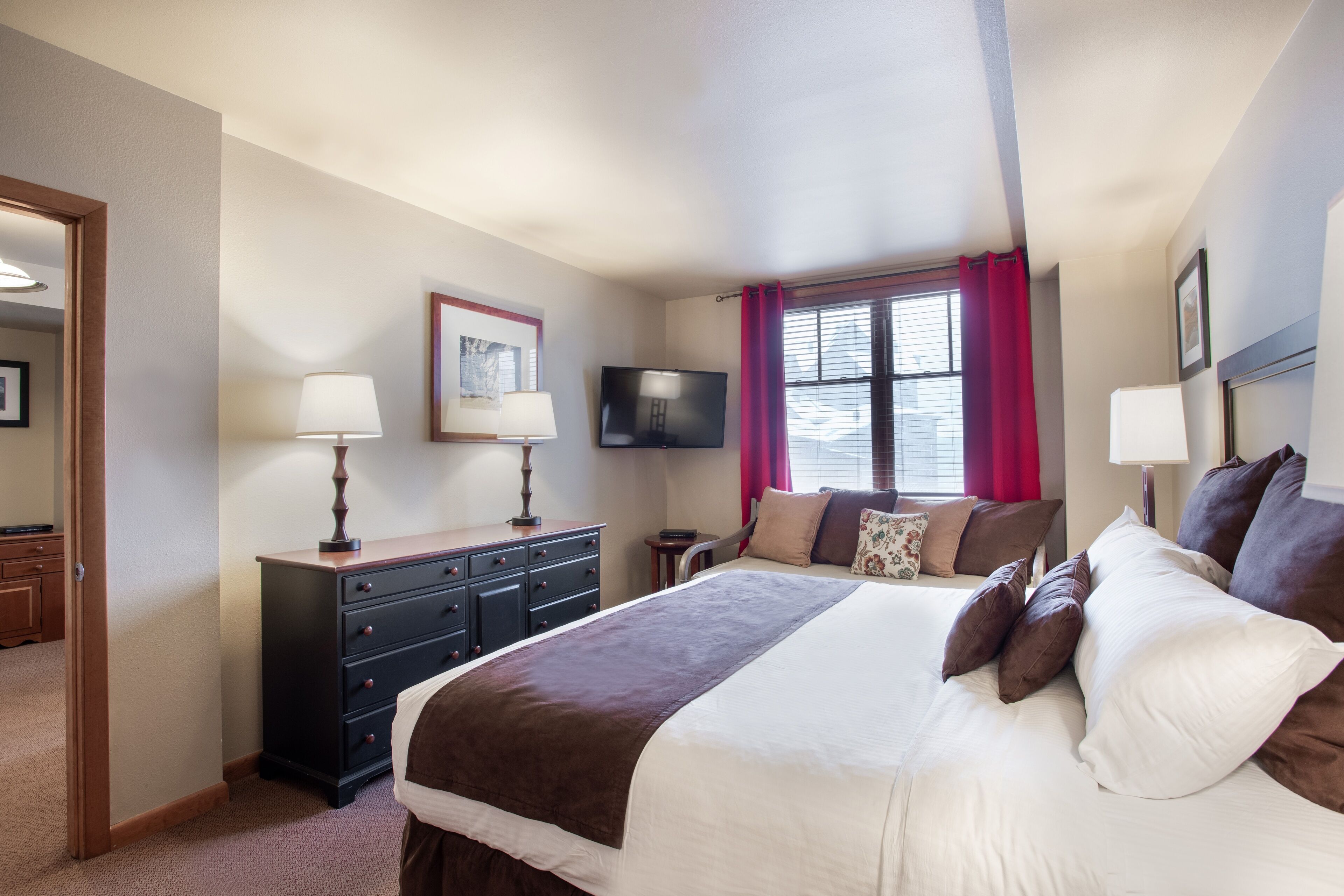 A hotel room with a large bed, a dresser, a TV, and a window with curtains.
