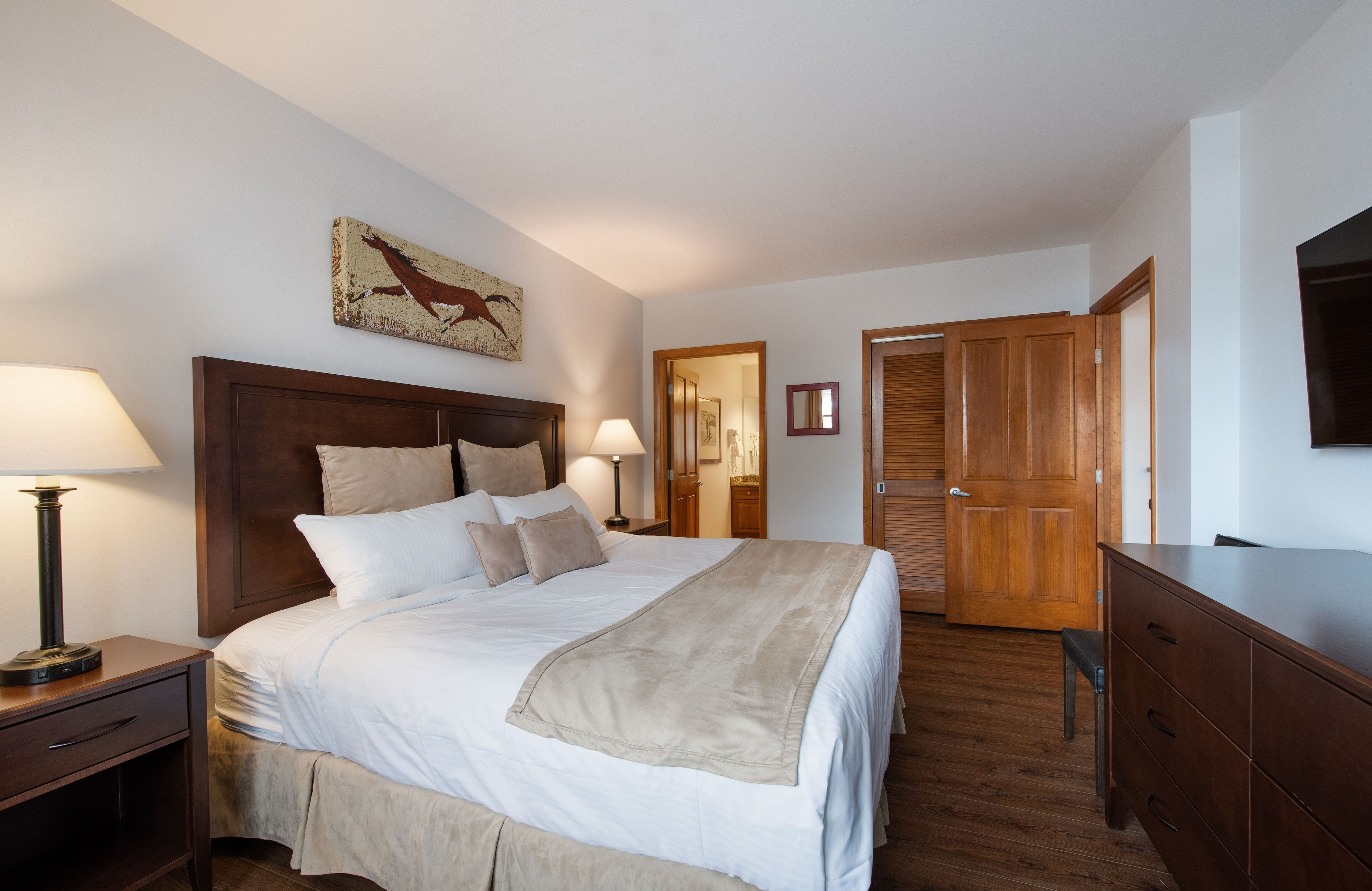 A neatly arranged bedroom with a large bed, bedside tables, a wooden wardrobe, a television, and a door leading to another room.