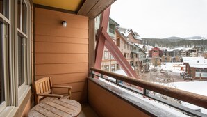 A balcony with a wooden table and chair, overlooking a snow-covered town with buildings and mountains in the background.