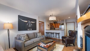 A living room with a sofa, coffee table, and a large painting of a bull. There is a kitchen area with stools and a bar.