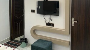 A modern bedroom with a TV mounted on the wall, a door, and a small stool. - Yatri Nivas By Sarvjeet Foundation (Varanasi)