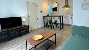 A modern living room with a TV, a coffee table, and a sofa. A kitchen is visible in the background with a microwave and bar stools. - SibsSanLúcar Lucero 2 (Sanlucar de Barrameda)