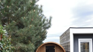 Une structure de sauna en bois, dotée d’une porte, qui est adjacente à un bâtiment muni d’une fenêtre.