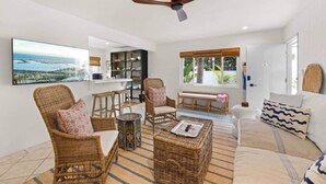 A spacious living room with wicker furniture, a ceiling fan, a large window with a view, and a kitchen area in the background. - Soul & Sea #1 – East Beach Guesthouse (Santa Barbara)
