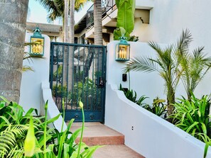 A gated entrance with a metal gate, a white wall, and lush green plants. - Soul & Sea #1 – East Beach Guesthouse (Santa Barbara)