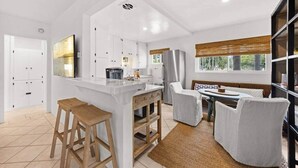 A modern kitchen with white cabinets, a central island with stools, a dining area with a table and chairs, and large windows with a view of trees. - Soul & Sea #1 – East Beach Guesthouse (Santa Barbara)