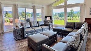A spacious living room with a large sectional sofa, ottomans, and a TV. Large windows offer a view of the outdoors. - Elevated Elegance with Golf Course Access & Panoramic Valley Views (Hamilton)