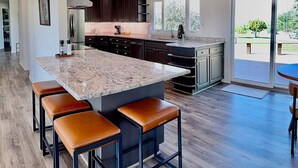 A modern kitchen with dark cabinets, a granite island, and a dining area with leather stools. - Elevated Elegance with Golf Course Access & Panoramic Valley Views (Hamilton)