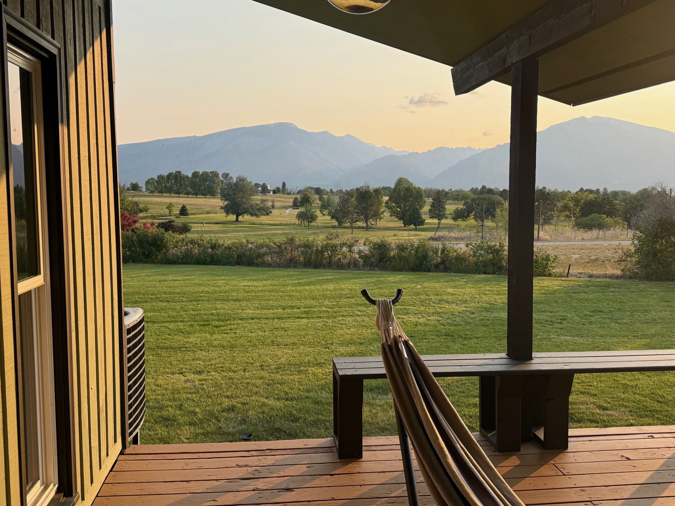 A wooden deck with a bench, overlooking a grassy field with trees and mountains in the distance.