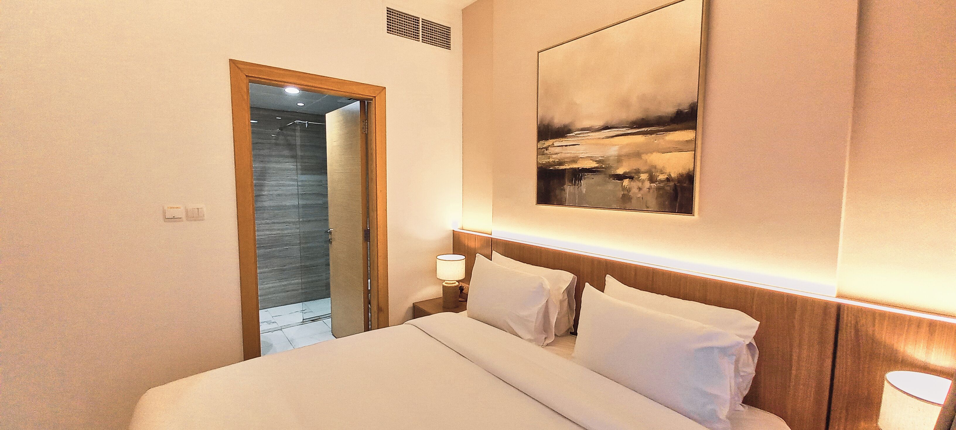 Une chambre d’hôtel avec un lit, une table de chevet avec une lampe, un tableau au mur et une porte donnant sur une salle de bain.