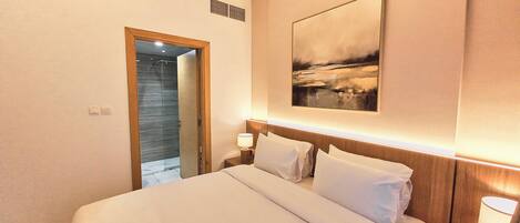 Une chambre d’hôtel avec un lit, une table de chevet avec une lampe, un tableau au mur et une porte donnant sur une salle de bain.