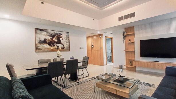 A modern living room with a dining area, a flat-screen TV mounted on the wall, and a large window. - Royal Home Deluxe Apartments, Beach View, 5min to Corniche (Abu Dhabi)