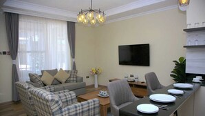 Un salon comprenant un canapé, des fauteuils, une table basse et une table à manger pour quatre personnes. Un téléviseur à écran plat est fixé au mur, et une plante est posée sur la table à manger.
