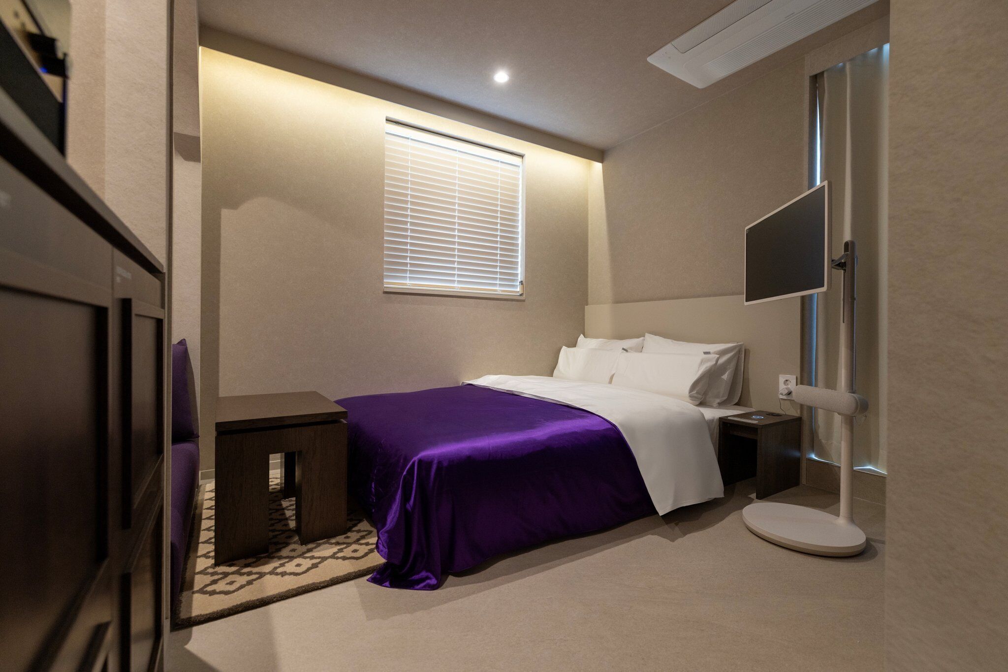 Un dormitorio con cama, mesita de noche, persianas en la ventana y televisor instalado en la pared.