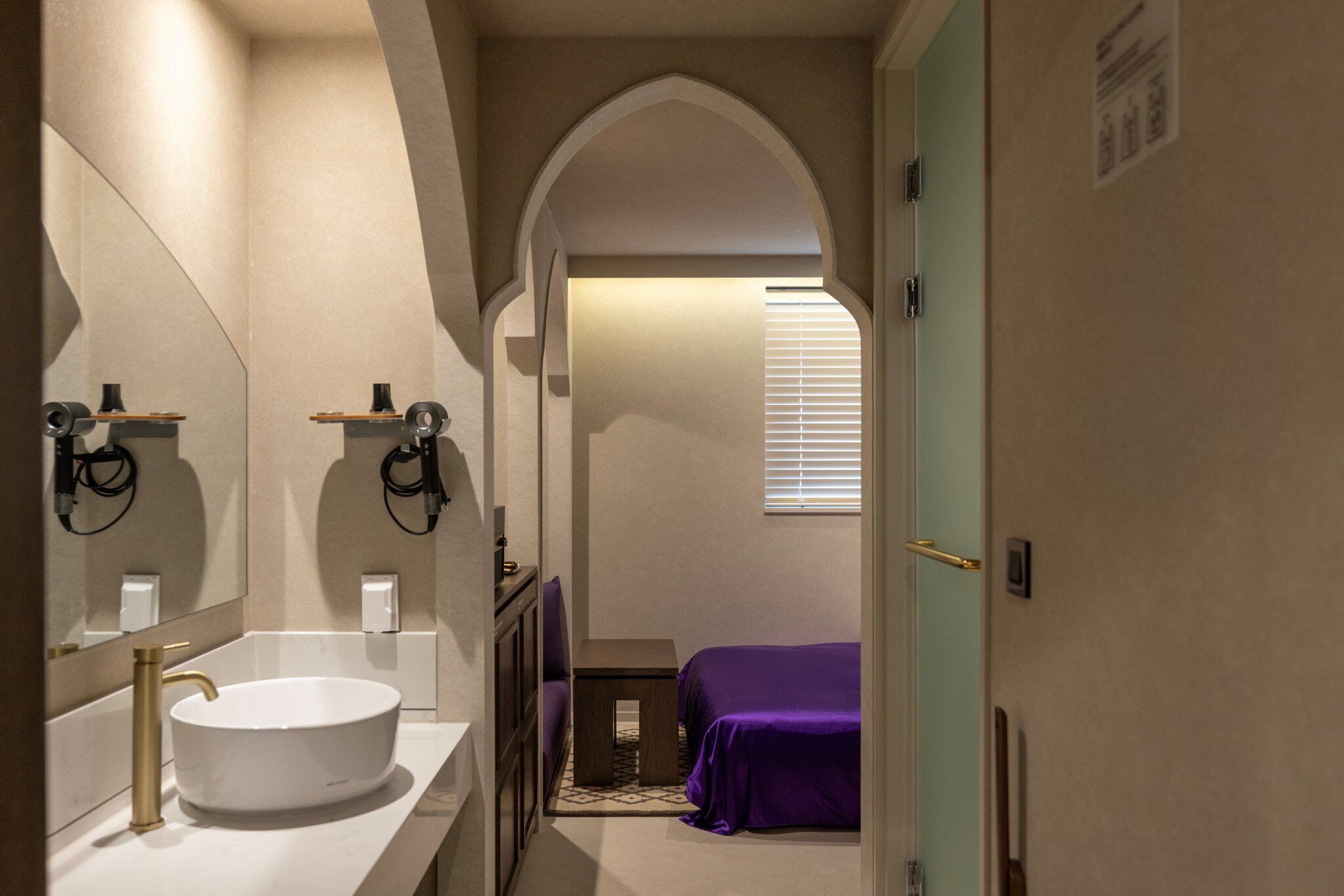 A bathroom with a white sink, a mirror, and a small table. There is a purple bed in the background.