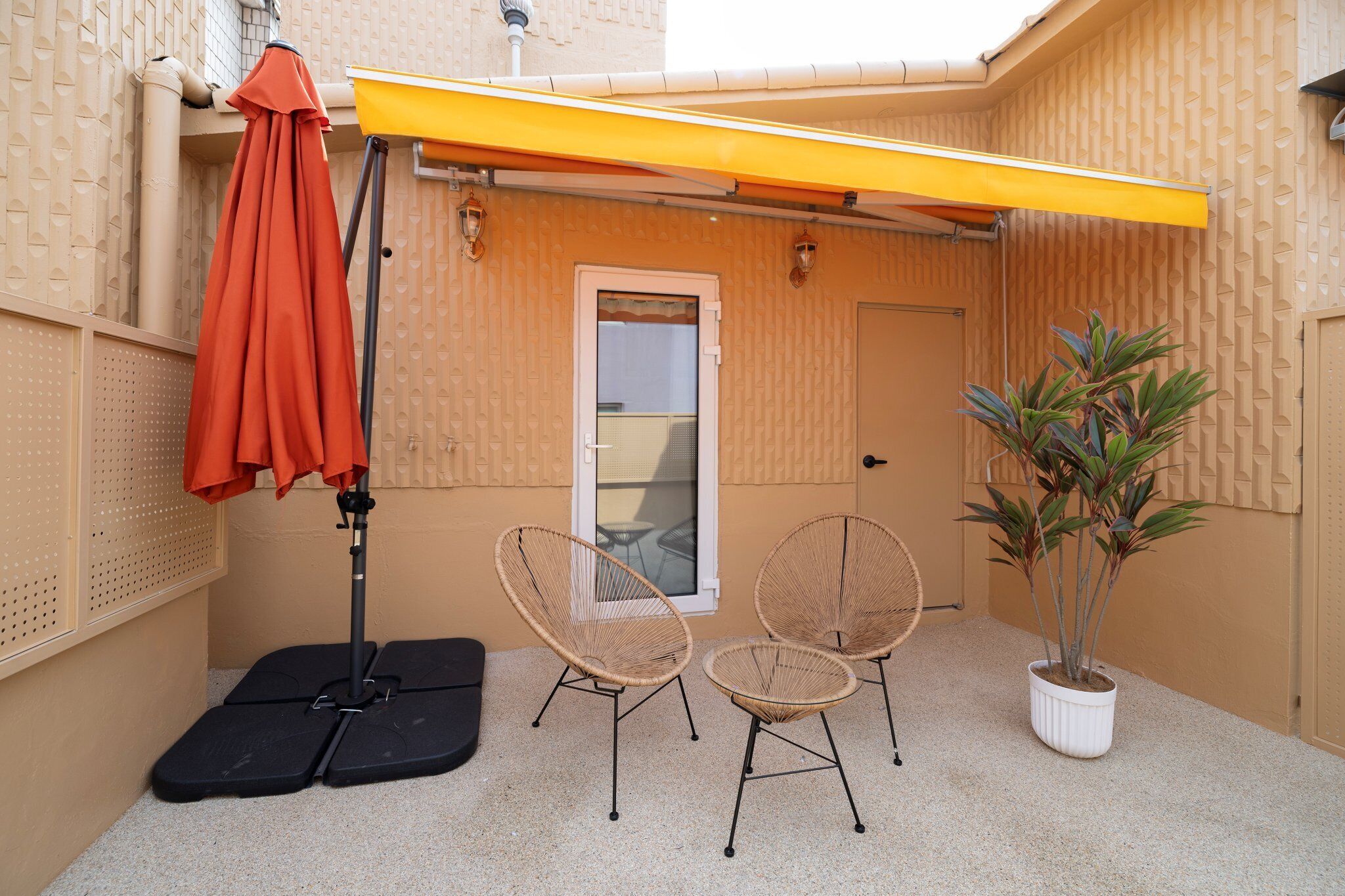 A small outdoor patio area with two wicker chairs, a potted plant, and a yellow awning.