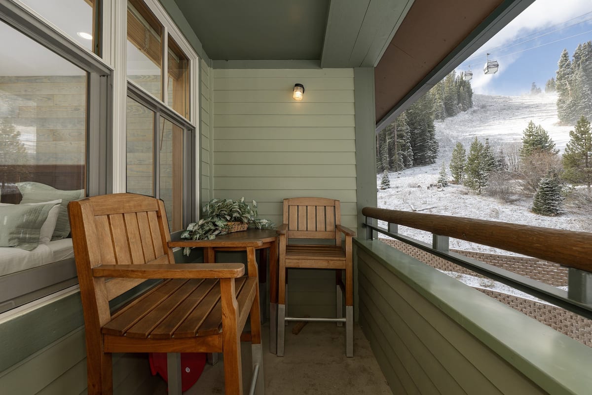 A balcony with wooden furniture, a small table, and a view of a snowy landscape with trees and a ski lift.
