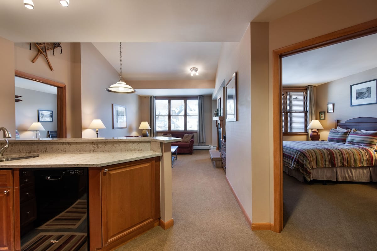 A spacious kitchen with wooden cabinets and a built-in dishwasher, a cozy living area with a sofa and a dining table, and a bedroom with a bed and a nightstand.