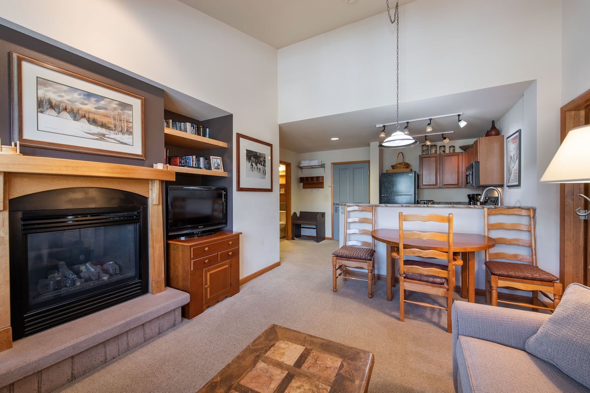 A living room with a fireplace, television, and wooden furniture. There is a kitchen area with a dining table and chairs.