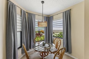 A corner room with a glass table, wicker chairs, grey curtains, and a hanging light fixture.