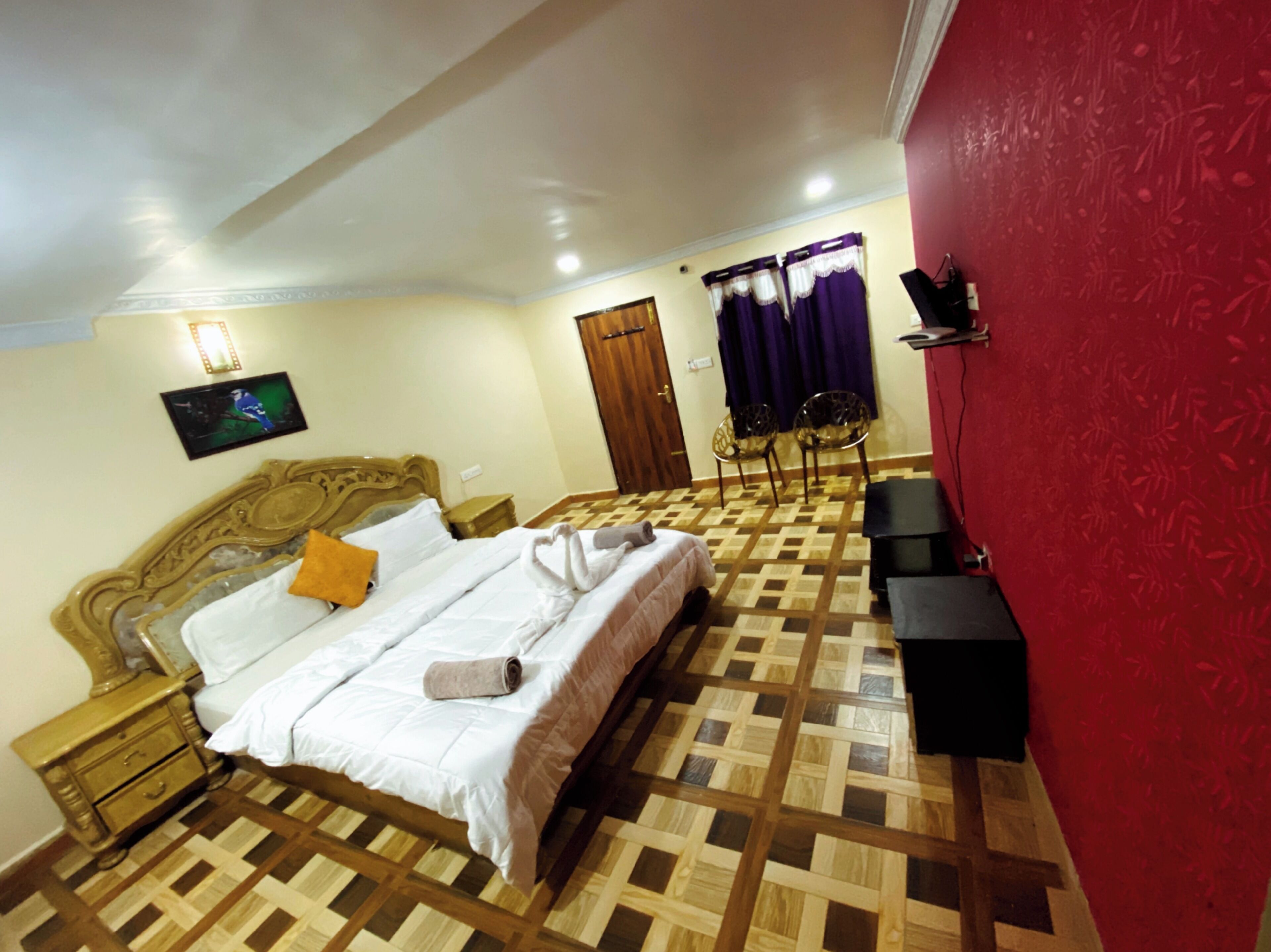 A bedroom with a bed, nightstands, a TV, a red wall, and patterned flooring.