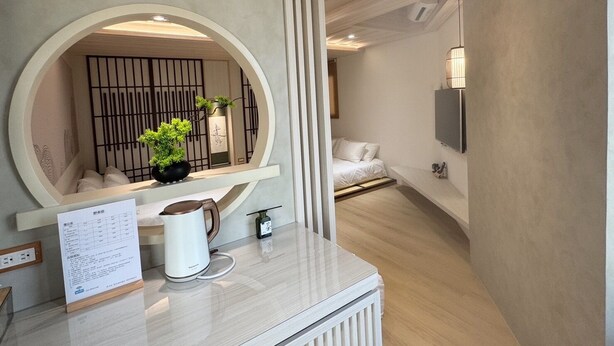 A hotel room with a large round mirror, a small potted plant, a white bed, and a flat-screen TV mounted on the wall. - yado B&B (Chaozhou)