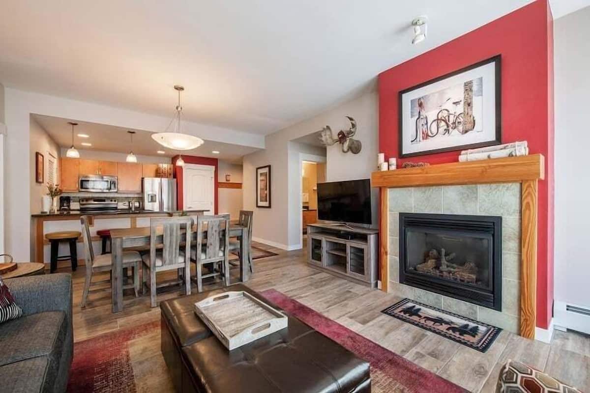 A modern living room with a fireplace, a dining area with wooden furniture, and a kitchen in the background.