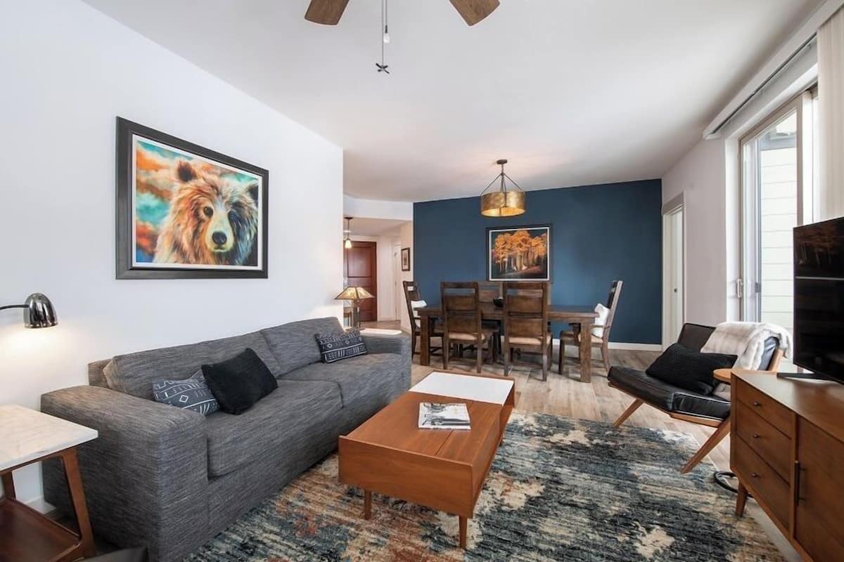 A modern living room with a grey sofa, coffee table, and a painting of a dog. There's a dining area with wooden chairs and a table, and a TV on a wooden cabinet.