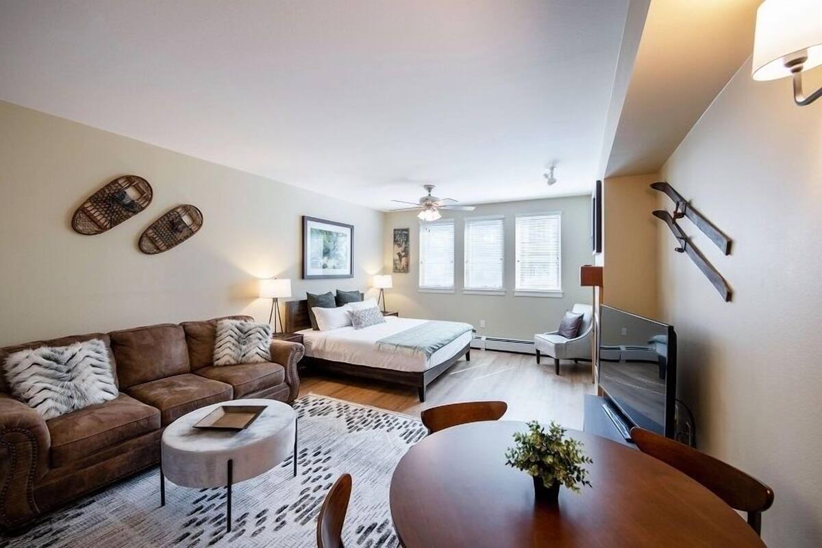 Un salon moderne avec un canapé, une table basse et une table à manger. On aperçoit un lit avec une tête de lit et un téléviseur au fond.