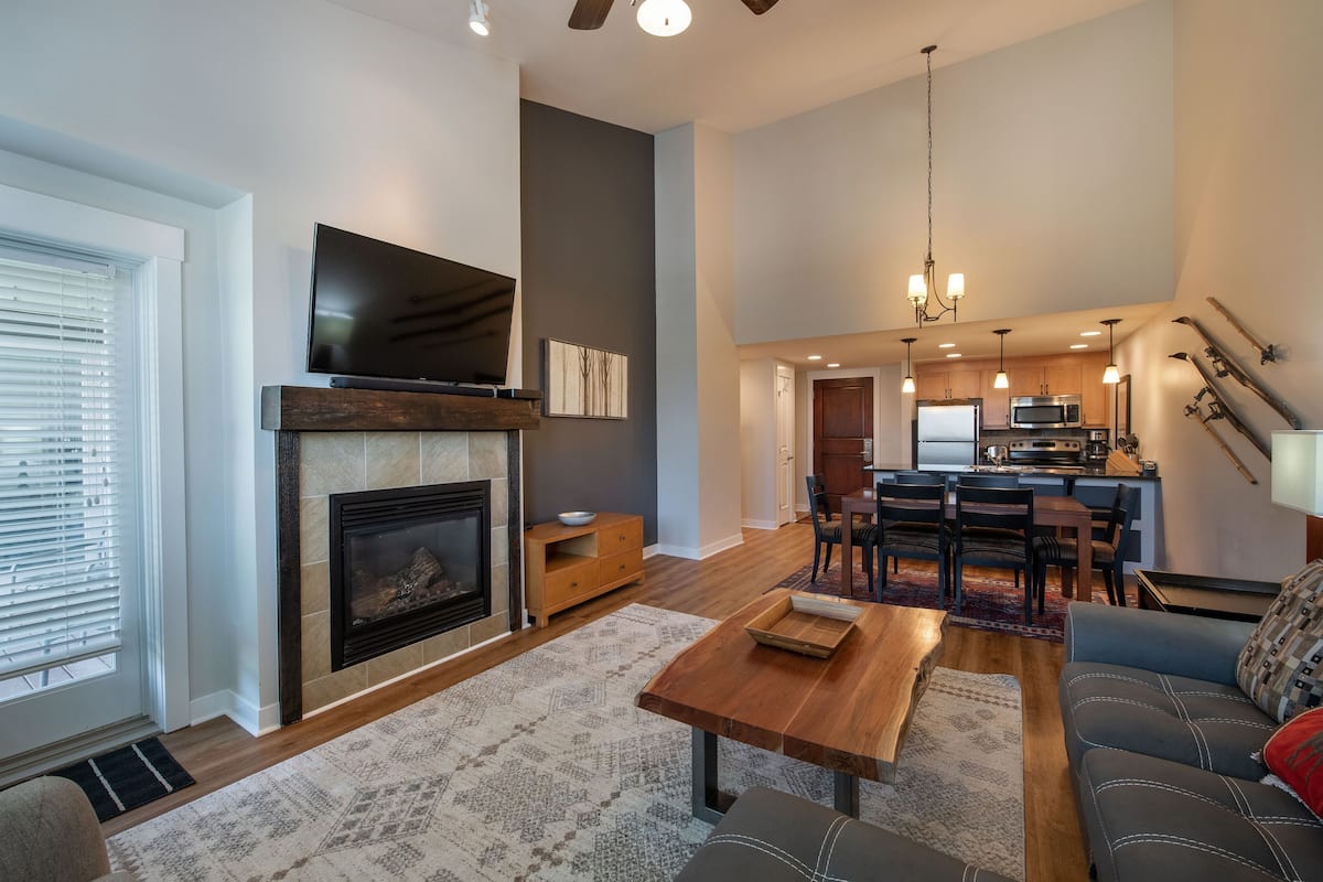 A spacious living room with a fireplace, a television, and a dining area with a kitchen in the background.