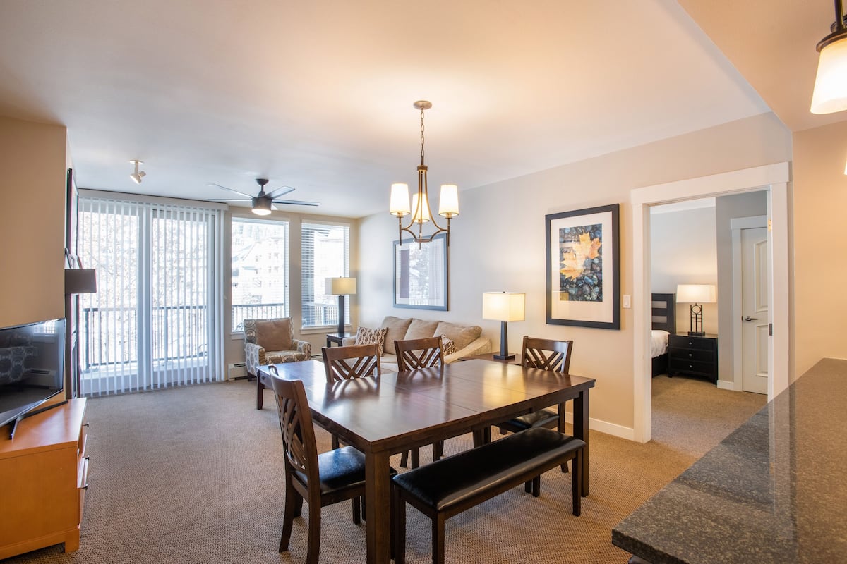 A spacious living room with a large dining table, comfortable seating, a ceiling fan, and a large window with blinds.