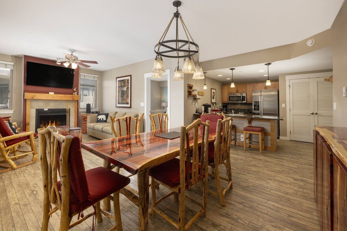 A spacious living area with a wooden dining table, red upholstered chairs, a fireplace, and a modern kitchen in the background.