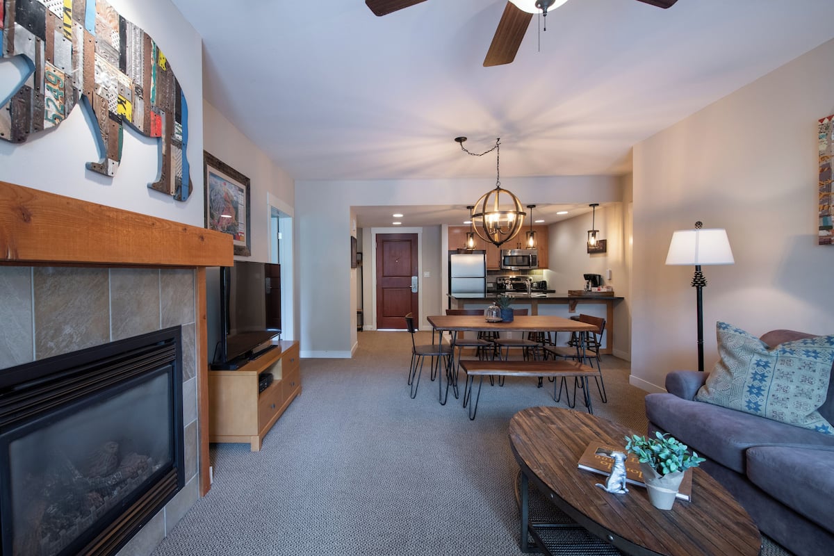 A modern living room with a fireplace, a dining area, and a kitchen in the background.