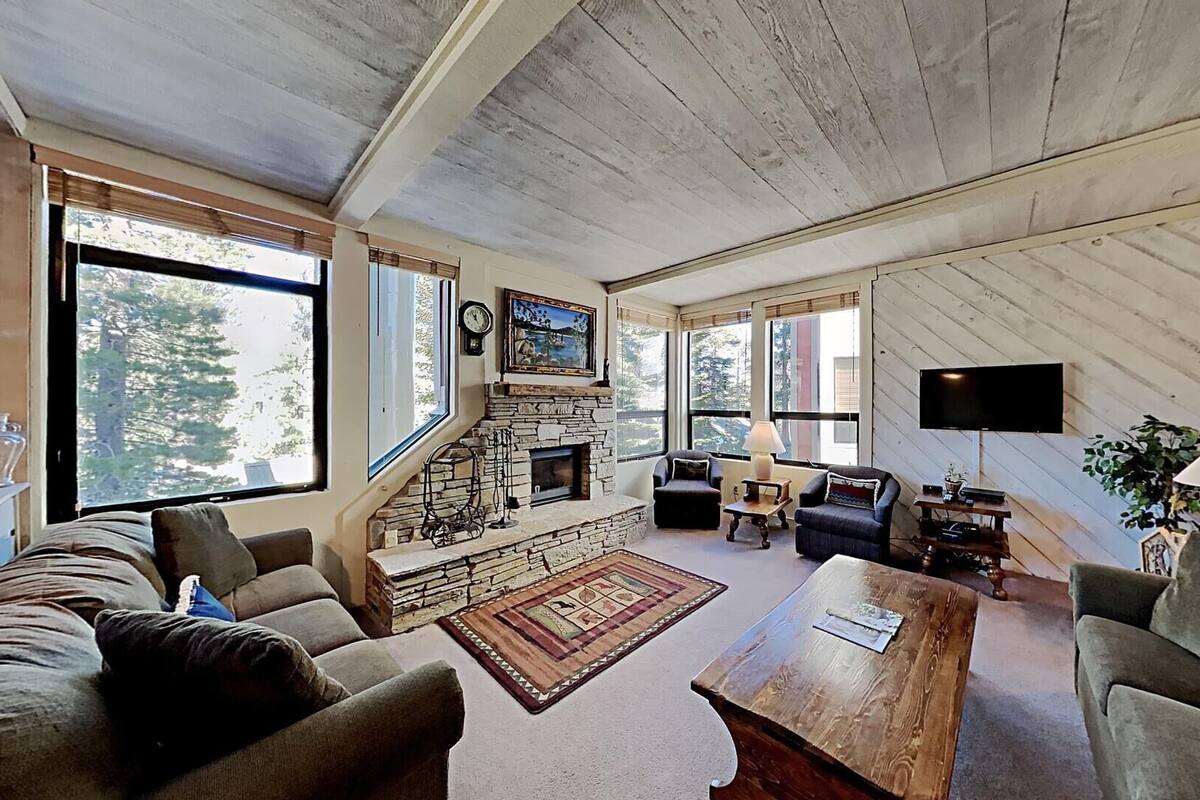 A cozy living room with a stone fireplace, comfortable seating, and large windows offering views of trees.