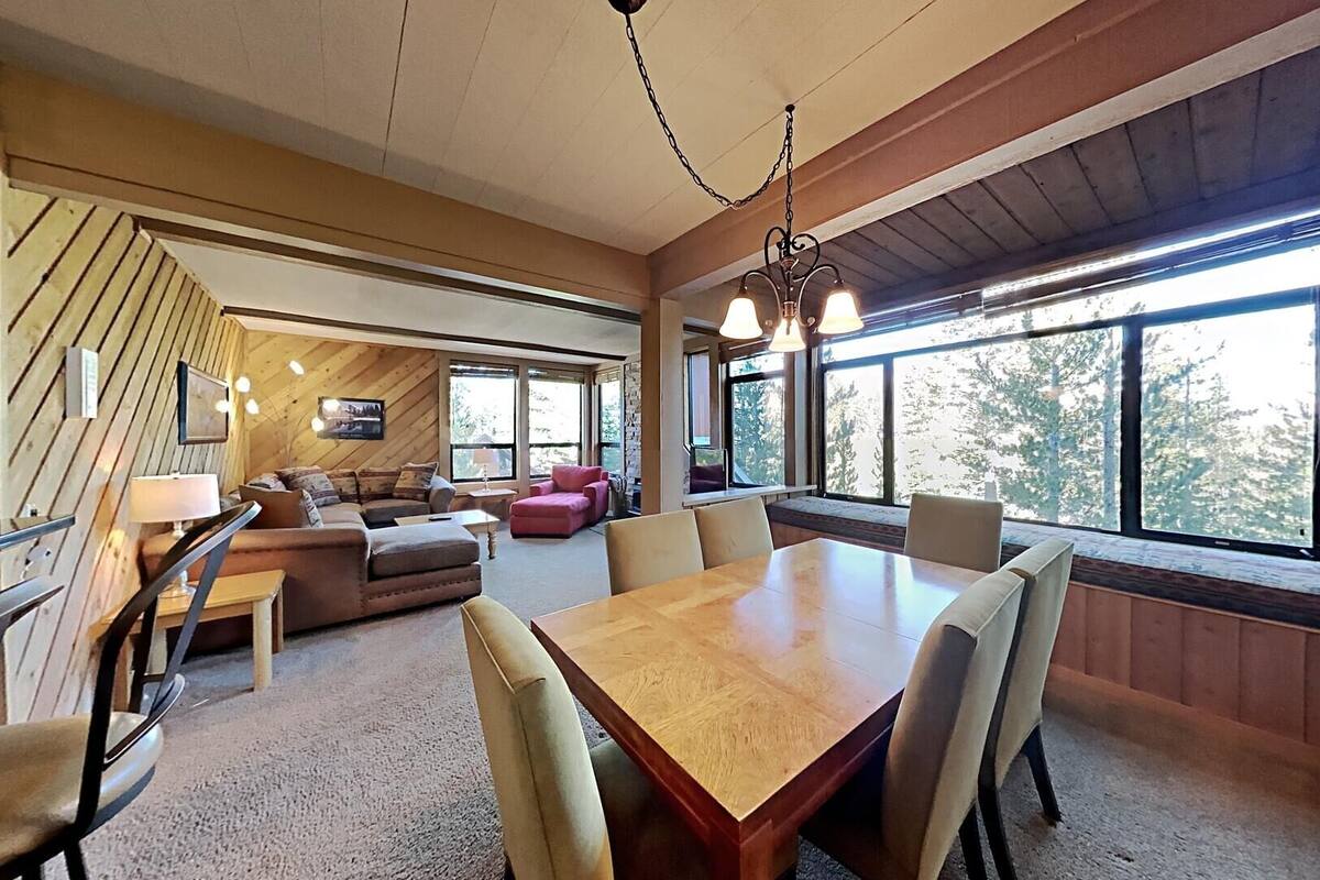 A spacious room with a large wooden dining table, comfortable seating, and large windows offering a view of the outdoors.