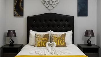 Una cama bien hecha con un cabecero negro, dos almohadas con la inscripción 'Cream BIG' y una disposición de toallas con forma de corazón sobre la cama.