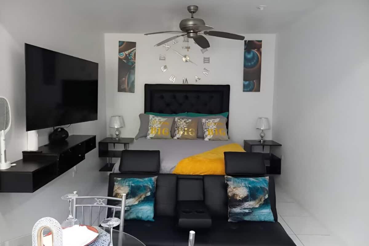 Una camera da letto moderna con una TV a schermo piatto, una ventola a soffitto, un letto con testiera imbottita e un comodino con lampada.