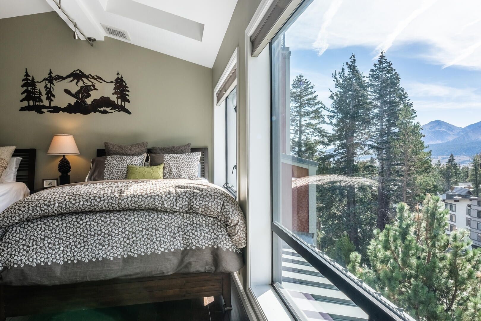 Ein Schlafzimmer mit einem Bett, Kissen, einer Lampe und einem großen Fenster mit Blick auf einen Wald und Berge.