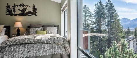 Ein Schlafzimmer mit einem Bett, Kissen, einer Lampe und einem großen Fenster mit Blick auf einen Wald und Berge.