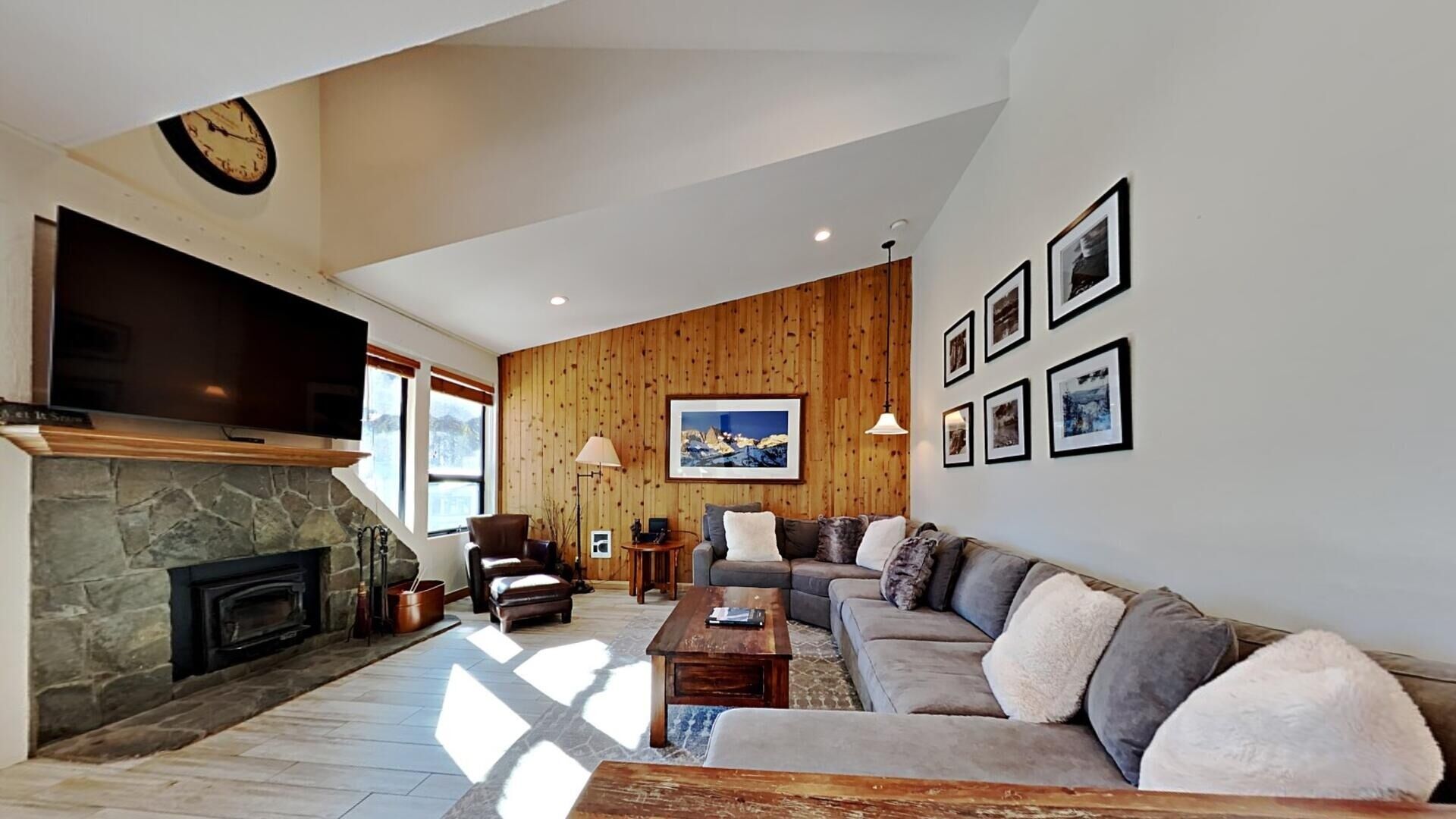 A living room with a stone fireplace, a large flat-screen TV, a wooden wall panel, a sofa, and framed pictures on the wall.
