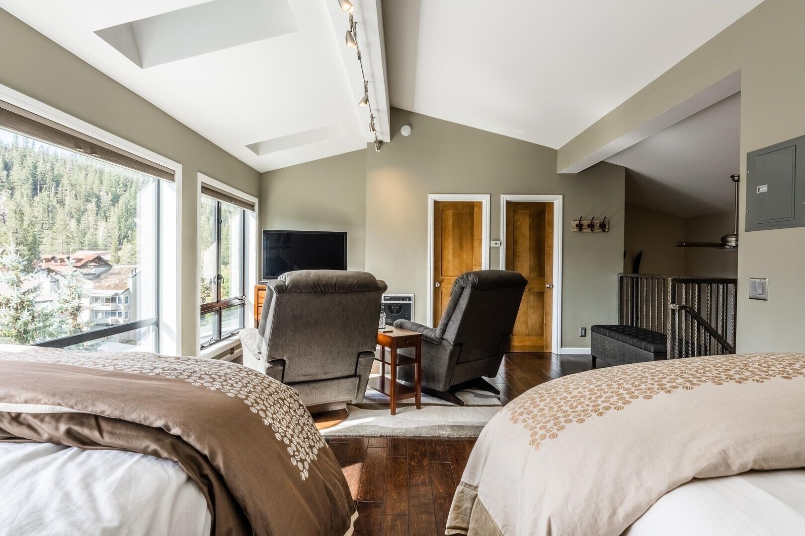 A bedroom with two beds, a desk, a chair, a TV, and a balcony with a view of houses and trees.