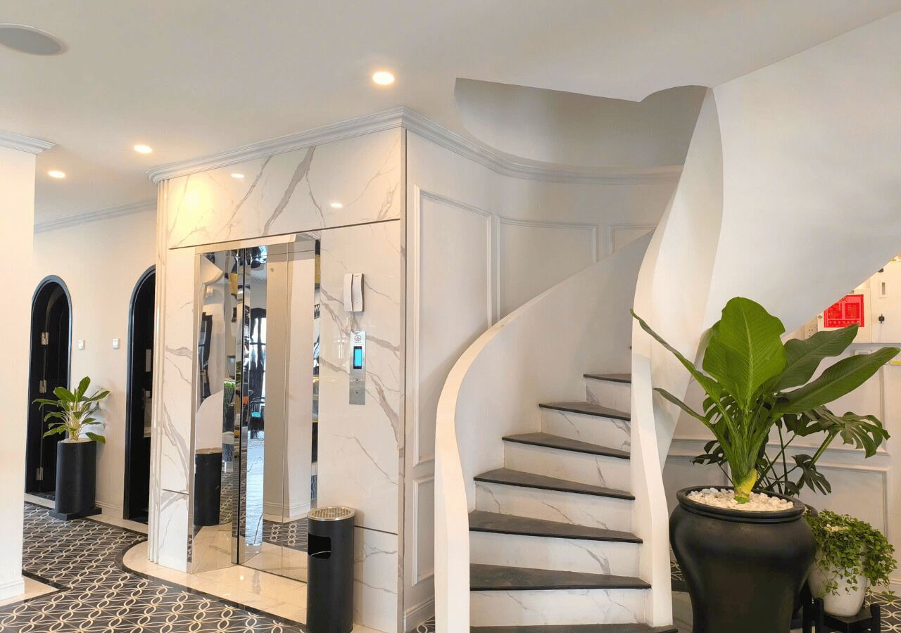 A modern interior with a marble staircase, a glass elevator, and potted plants.