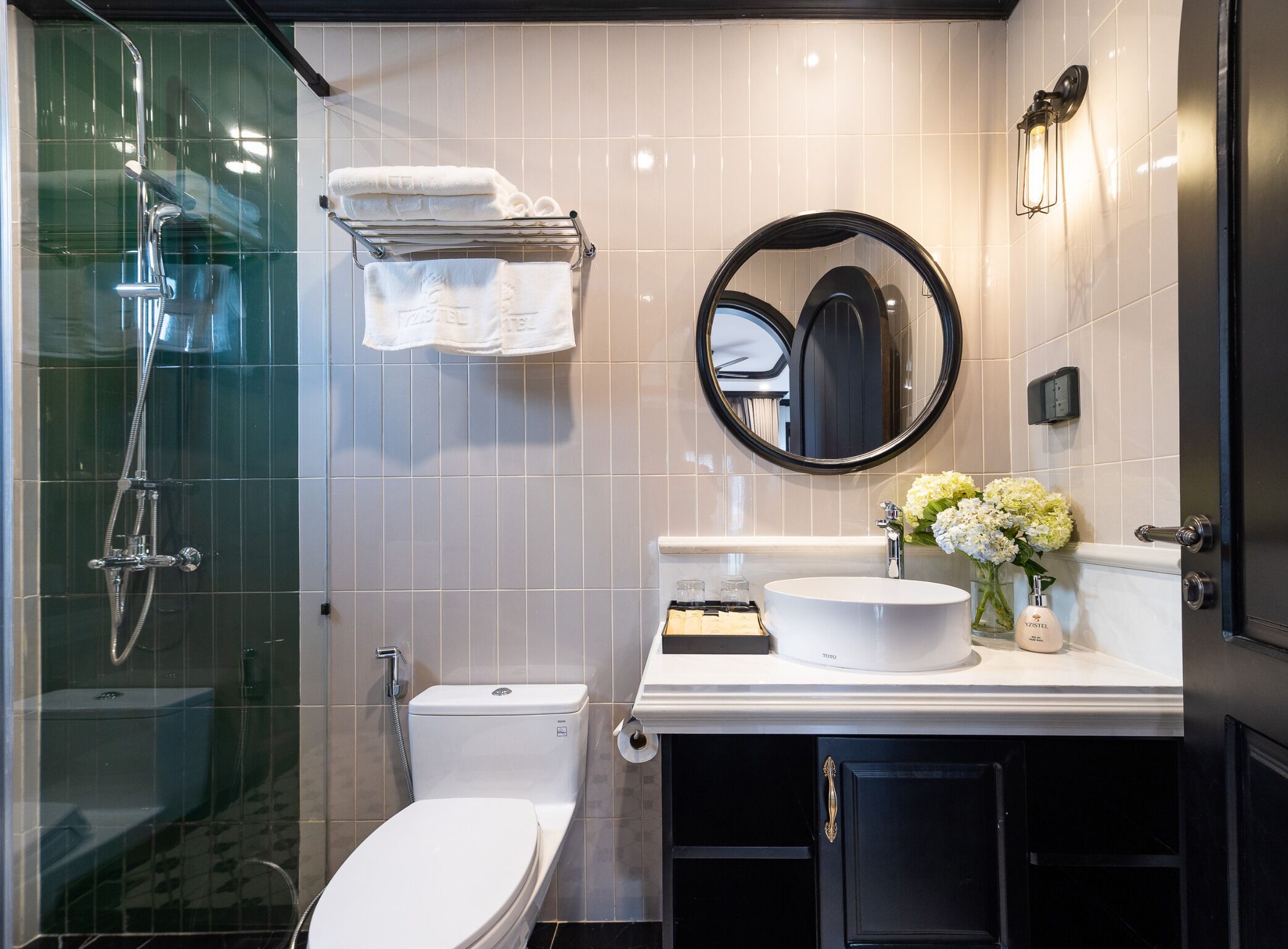 Un baño moderno con ducha envuelta en vidrio, inodoro blanco, tocador con espejo redondo y lavabo con un florero.