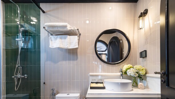 A modern bathroom with a glass-enclosed shower, a white toilet, a vanity with a round mirror, and a sink with a vase of flowers. - Yzistel Hoi An (Da Nang)