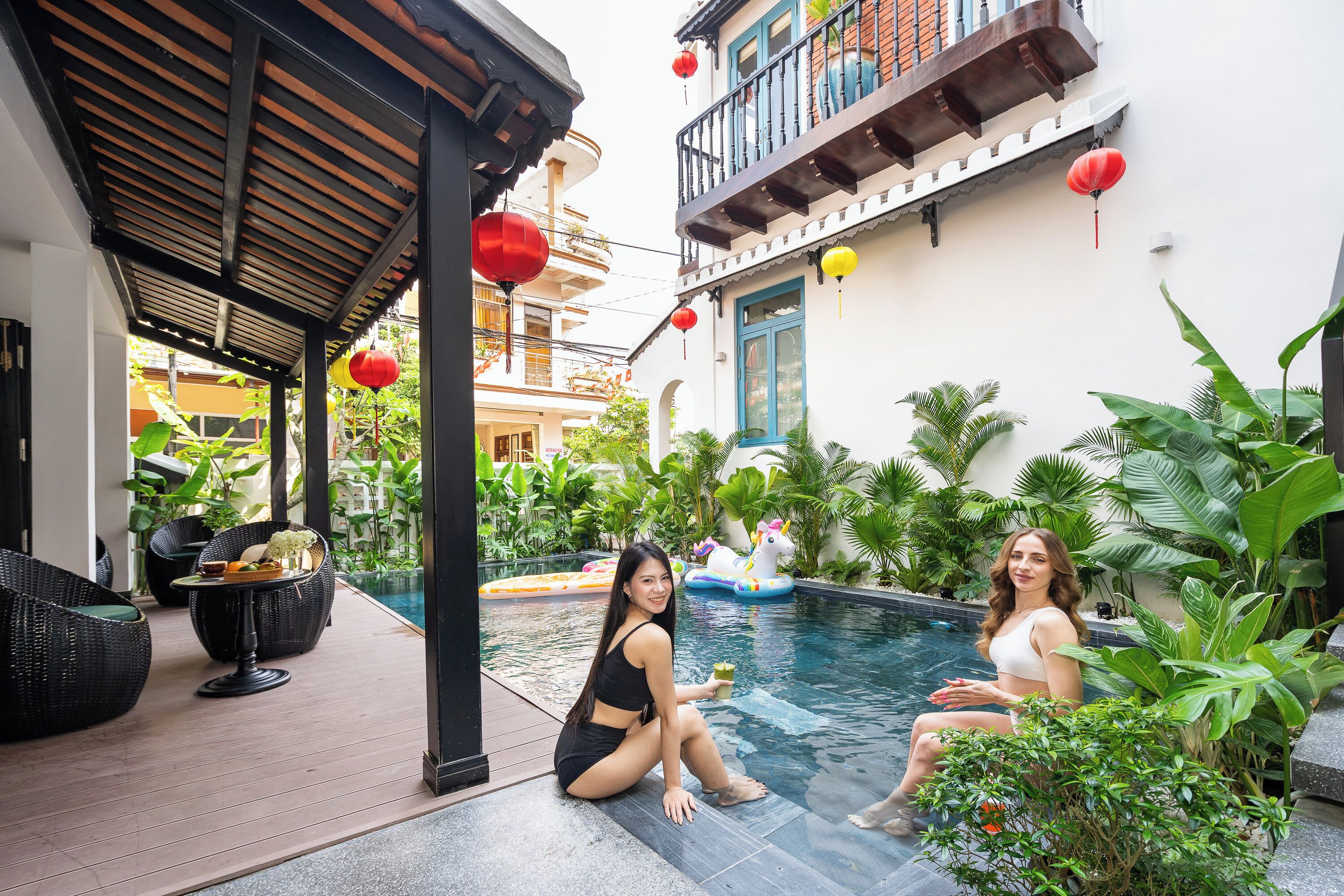 A poolside area with a covered seating area, a pool, and surrounding greenery.