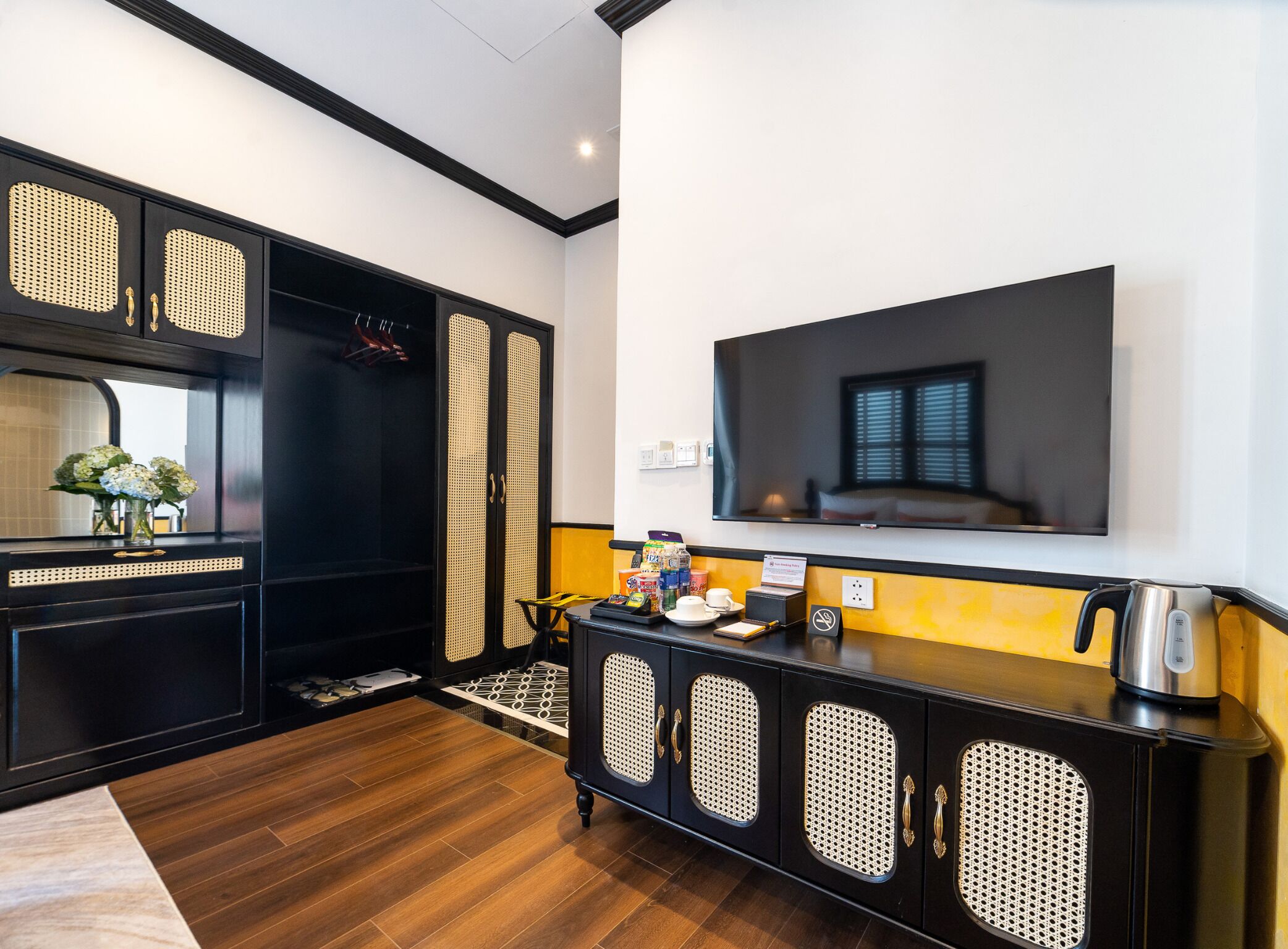 Een moderne hotelkamer met een grote flatscreen-tv aan de muur, een zwart en geel console tafeltje met waterkoker en theeset, en een houten vloer.