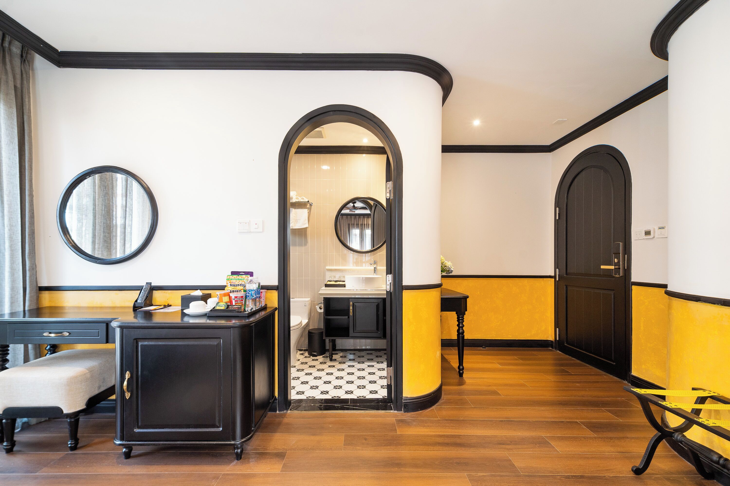 A hotel room with a desk, chair, and a small table with a mirror. There is a door leading to a bathroom with a sink and toilet.