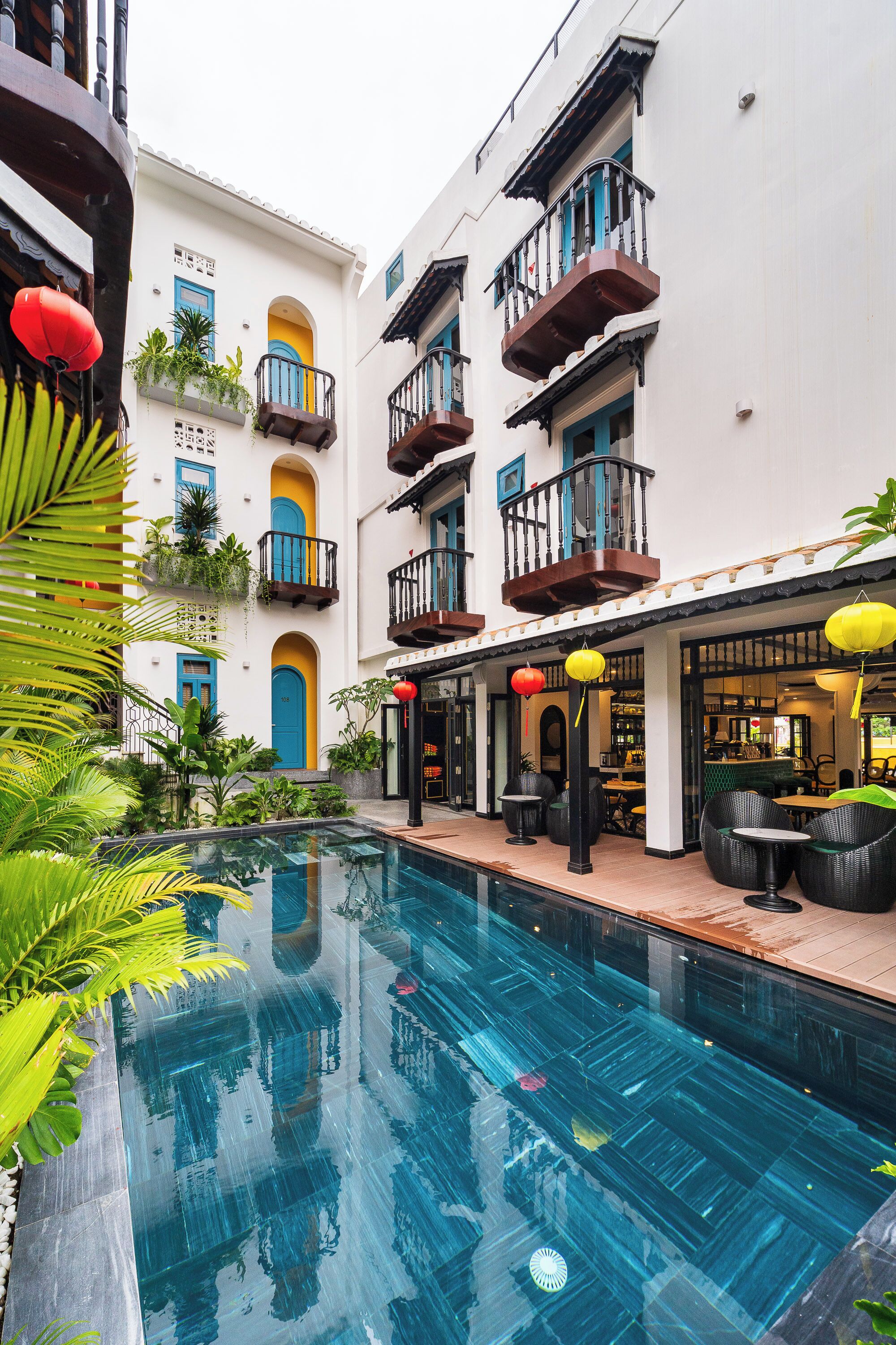 A pool area with a clear blue pool, surrounded by lush greenery and a building with balconies and outdoor seating.
